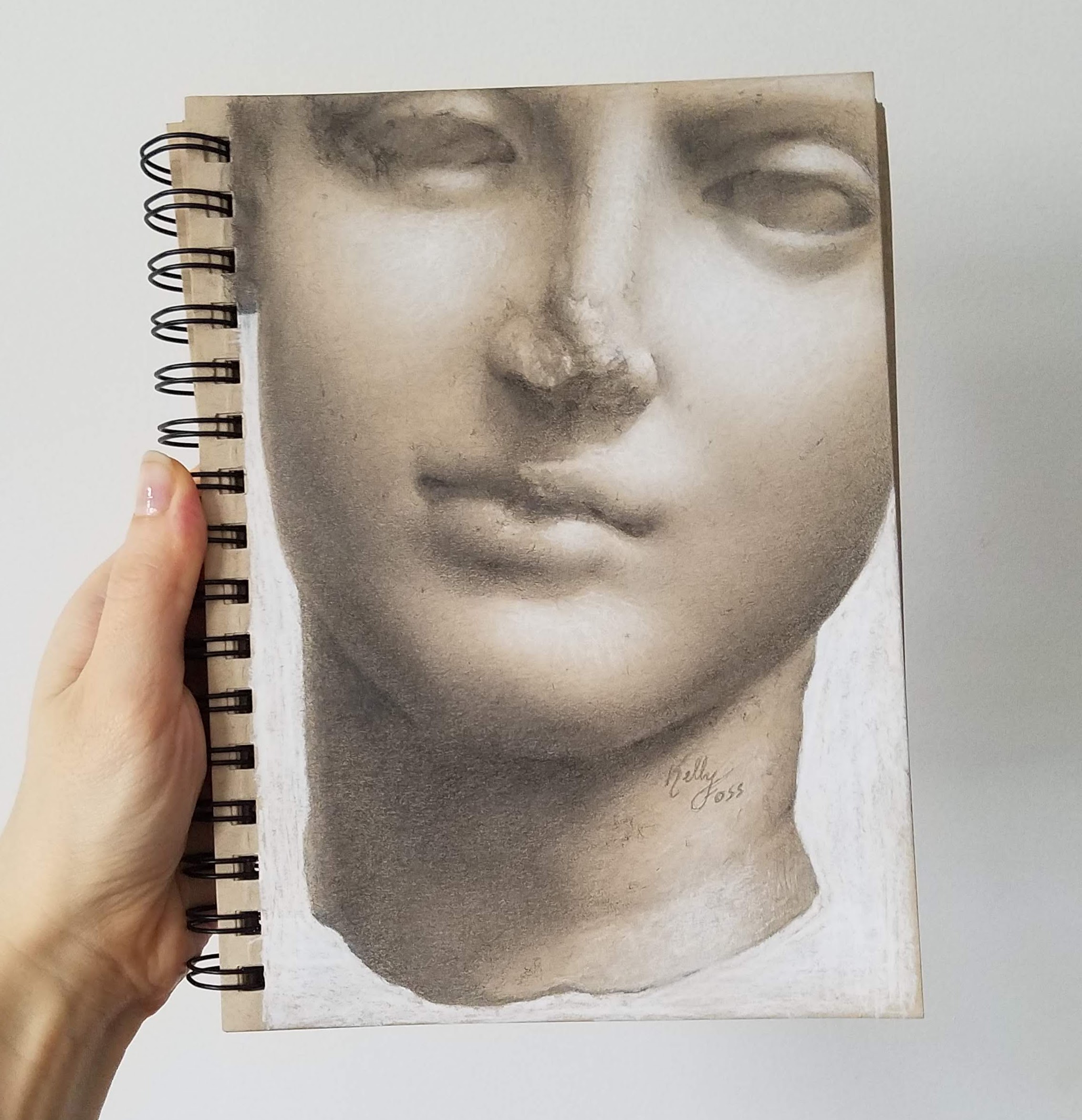 Handholding a sketchbook featuring a detailed gray-scale drawing of a sculpture's face.