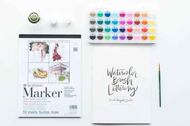 "Art supplies including watercolor paints, markers, and brush lettering book."